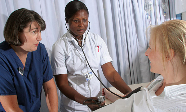 Nursing student takes patient's pulse at hospital bedside, with support of nurse standing beside her