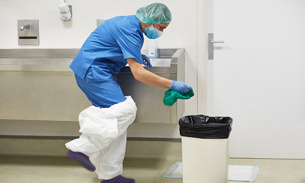 A nurse in uniform in a clinical setting removing PPE, including gloves and trousers, and disposing of them in a bin