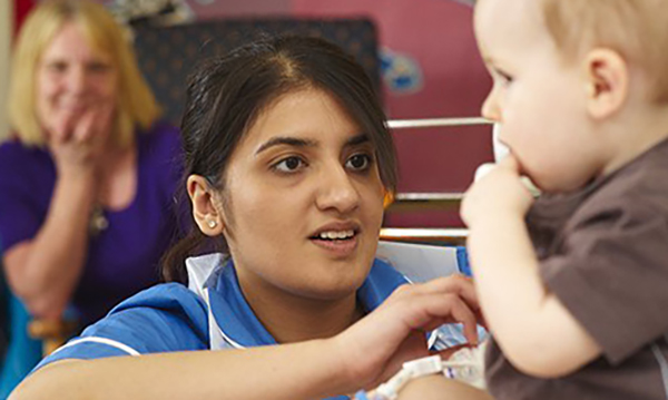 A nuse smiles at child as mother looks on. 29% of children in England are living in poverty, fewer are being immunised and there are growing health inequalities with more of the poorest becoming obese