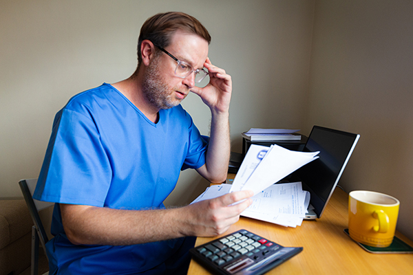 Nurses’ pay and pension worries: concerned nurse sits at desk, head in hand as he read documents that could be bills or bank statements