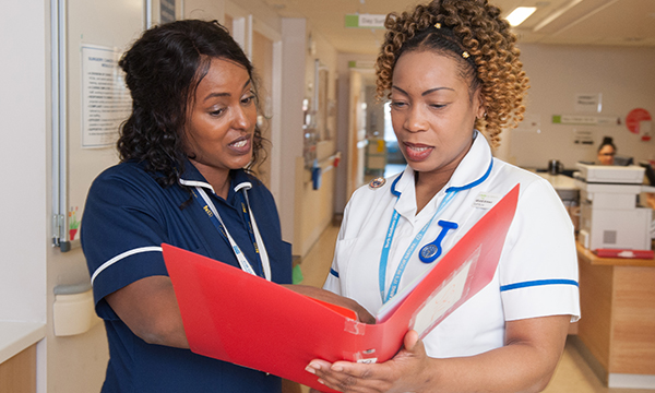 A newly-qualified nurse shares a file binder of notes with a senior nurse on a hospital ward 