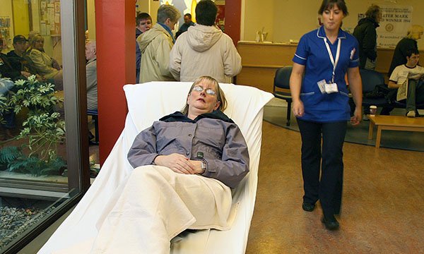 woman lies on a hospital trolley in front of a window in a hospital reception area