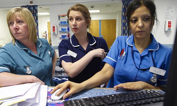 Three nurses stand together at ward station. They appear busy