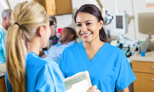 A manager chats with a nurse in a hospital ward, while behind them a patient in a hospital bed is attended by other nurses