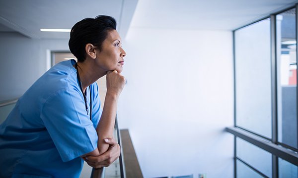 NHS nurse redundancy: pensive nurse leans on a corridor railing as she gazes out of a window